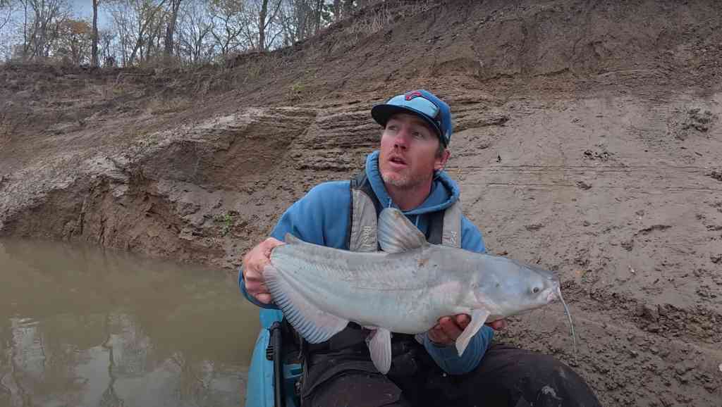 Spencer Bauer holds a solid blue catfish while seated in his kayak against a muddy shoreline.